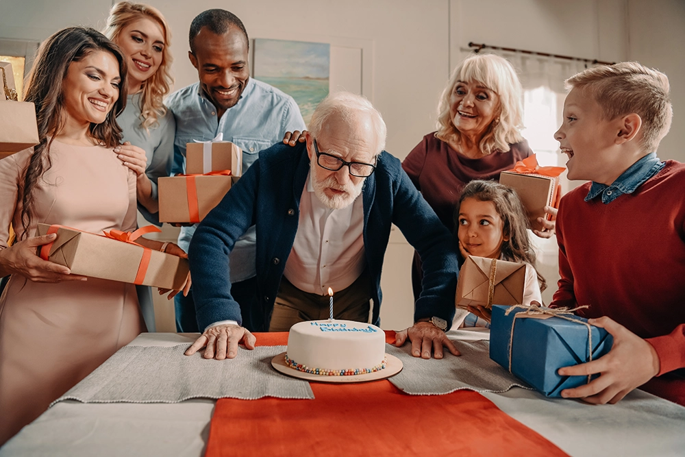 Elderly man blowing out birthday candles on a cake with family surrounding him.