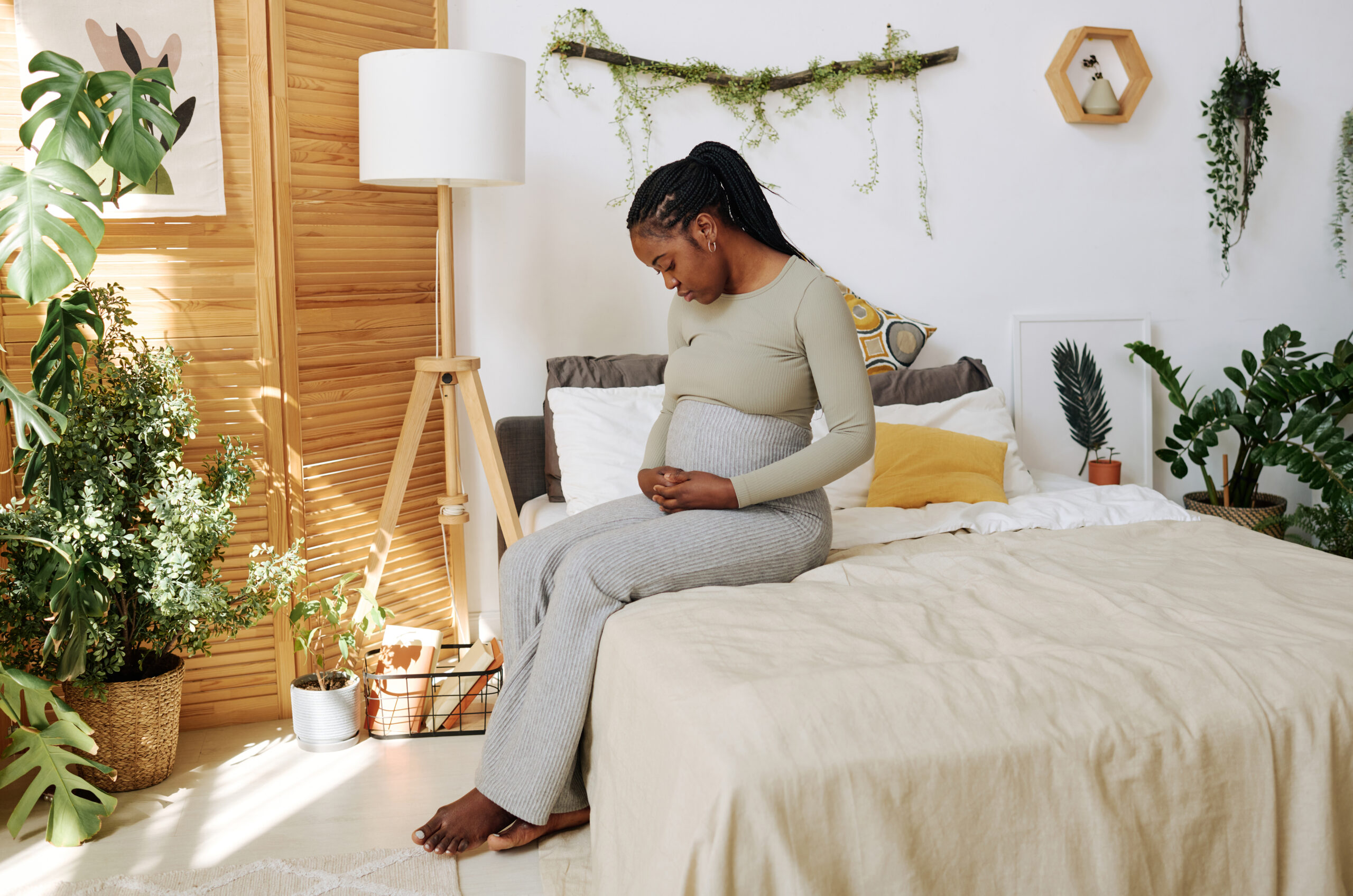 African sad woman looking at her pregnant belly while she sitting on bed in bedroom